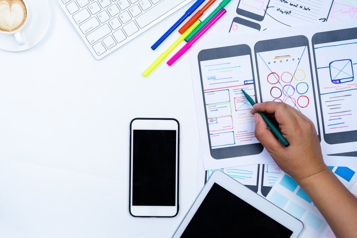 A person sketches a mobile app wireframe on paper for senior living websites, with a smartphone, tablet, colored pens, keyboard, and coffee cup on a white desk.