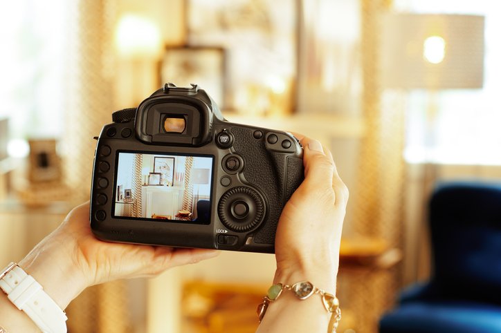 A person holds a digital camera, photographing a well-lit, decorated interior room—perfect for showcasing on a senior living website—visible on the camera's display screen.