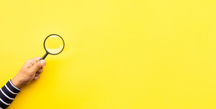 A hand holding a magnifying glass over a plain yellow background, symbolizing the search for information on a senior living website.