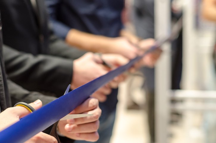 Several people hold scissors and cut a blue ribbon during a senior living grand opening ceremony, symbolizing the launch of a new facility or event.