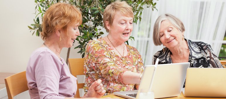 Three older women sit at a table using laptops together, discussing and collaborating as one gestures toward another’s screen—perhaps exploring a senior living website for helpful resources.