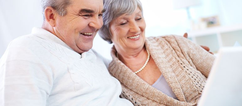 An older man and woman sit close together on a couch, smiling while looking at a laptop screen—exploring senior living marketing options online.