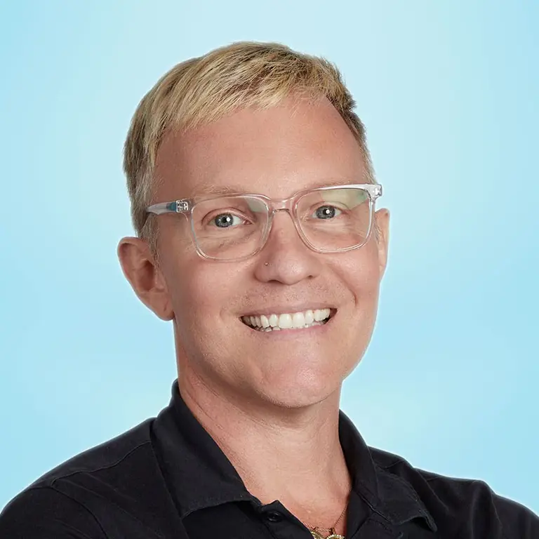 A person with short blond hair, clear glasses, and a black collared shirt smiles at the camera against a light blue background.