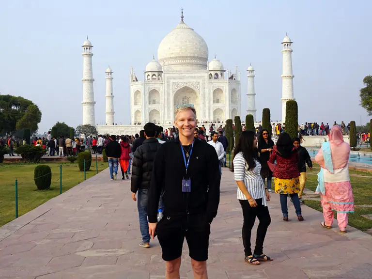 A person stands smiling in front of the Taj Mahal, surrounded by a crowd of tourists on a bright day.