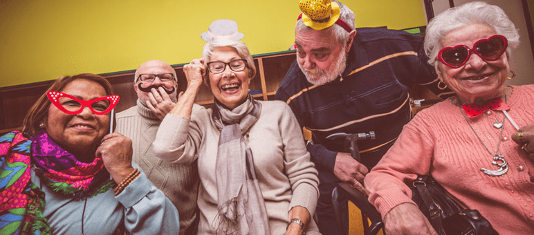 Five older adults wearing festive glasses, hats, and accessories, smiling and posing together at a senior living marketing event indoors against a yellow background.