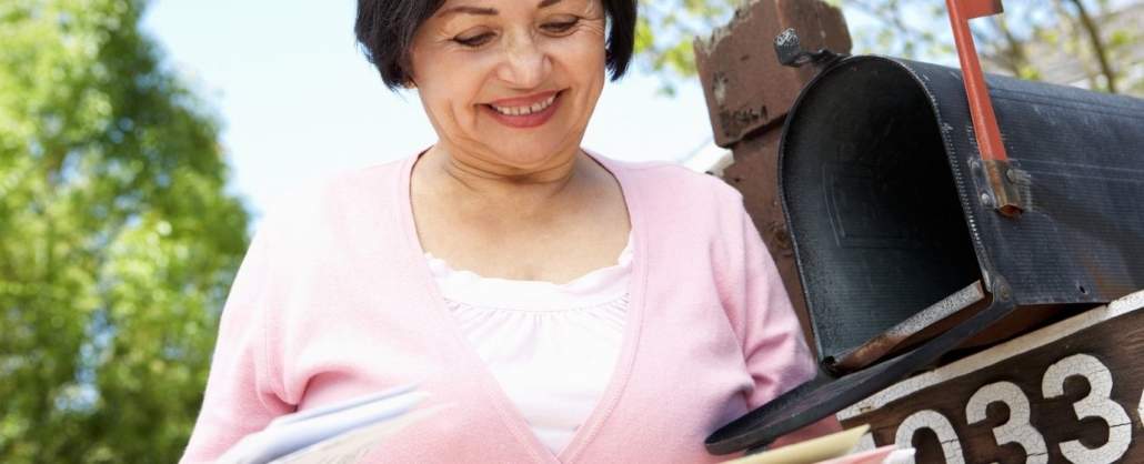 A woman stands by an open mailbox, holding and looking at a stack of mail while smiling—an uplifting moment that captures the personal touch of senior living marketing.