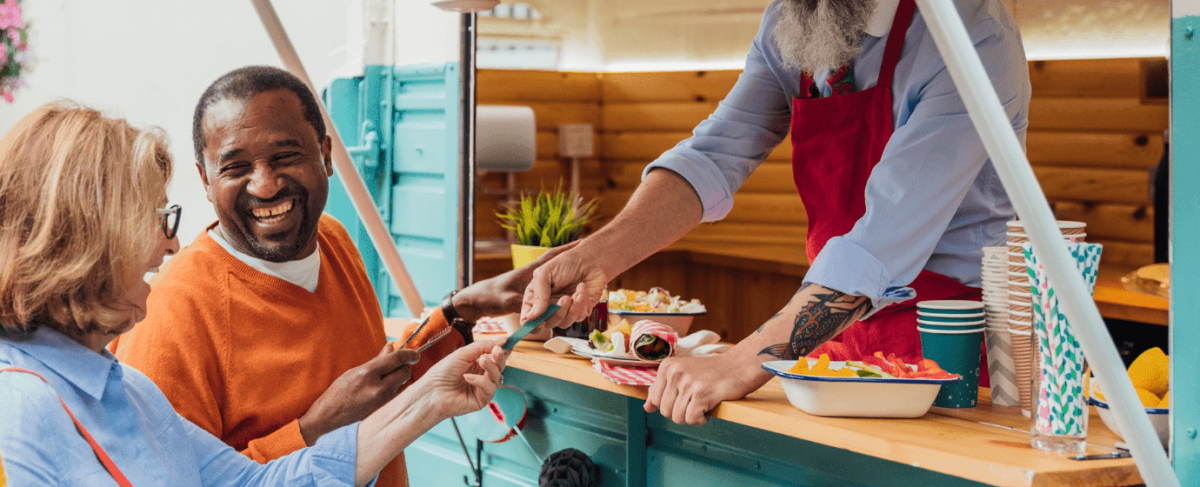 Two customers smile and pay at an outdoor food truck during a lively senior living marketing event, as a vendor in a red apron hands over food and drinks.